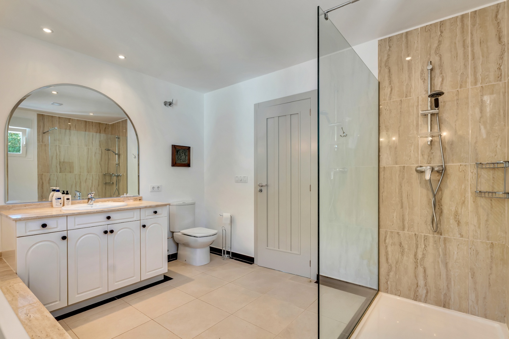 Elegant bathroom featuring a walk-in shower with natural stone tiling, a white vanity with marble countertop, and a large arched mirror under recessed lighting.