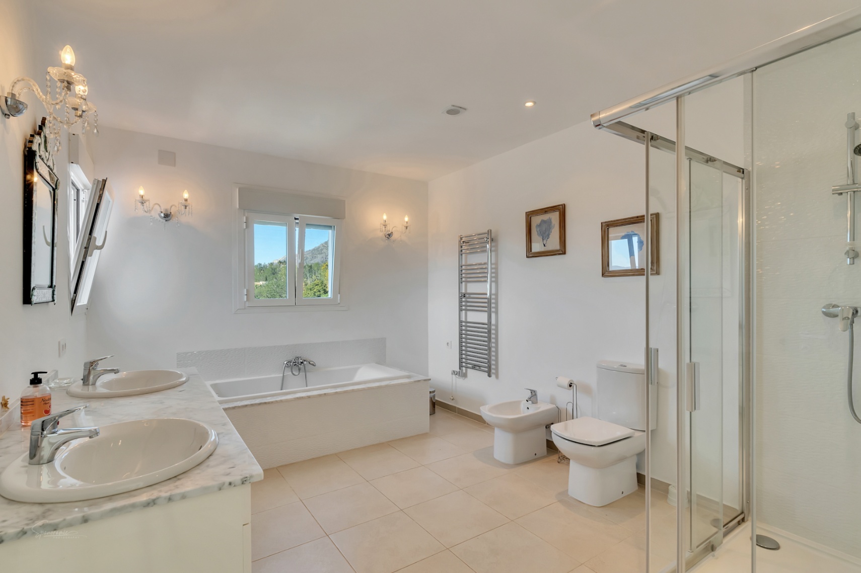 En-suite bathroom featuring a marble double vanity, built-in bathtub with mountain views, walk-in glass shower, and classic wall sconces over neutral tiled flooring.