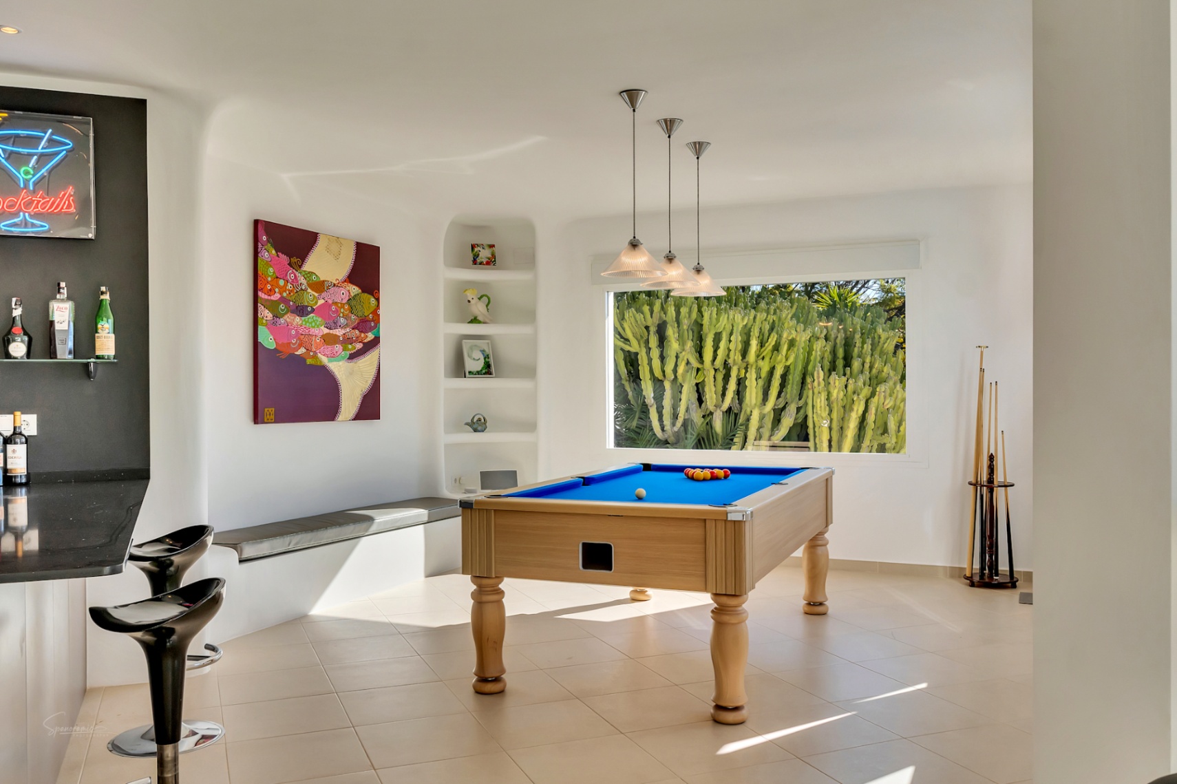 Entertainment room featuring a professional pool table, an integrated bar area with neon signage, and a large window framing garden cacti against smooth white Ibiza-style walls.