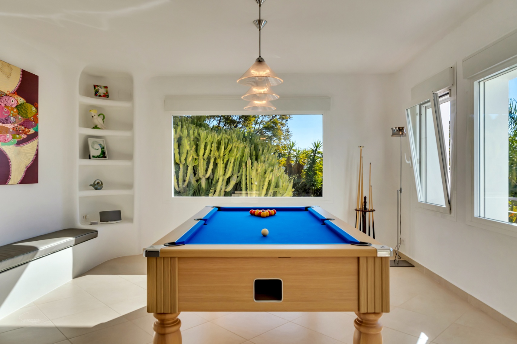 Entertainment room featuring a wooden pool table with blue felt, built-in shelving, and a large window overlooking a lush cactus garden.