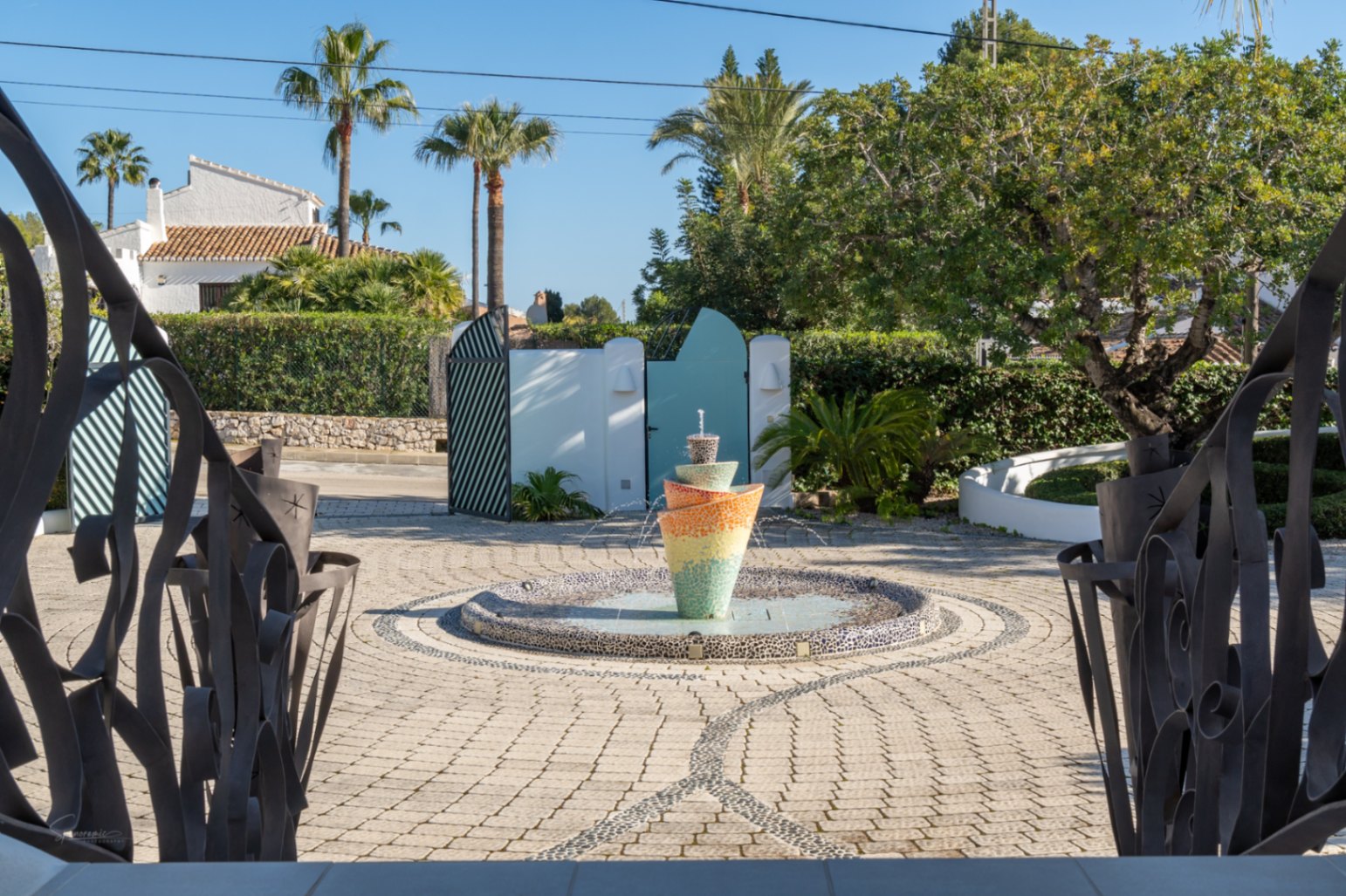 Entrance view through decorative iron railings showcasing a paved courtyard with a central mosaic fountain, palm trees, and a secure perimeter wall.