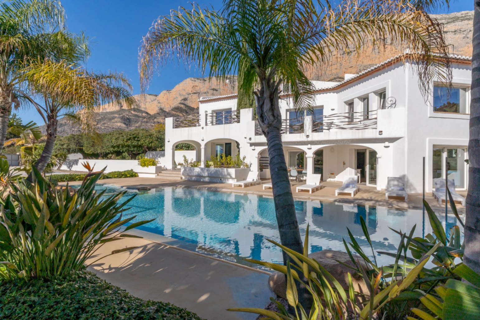 Exterior view of the Ibiza-style villa featuring a large swimming pool, arched ground-floor porches, and upper terraces set against the Montgó mountain.