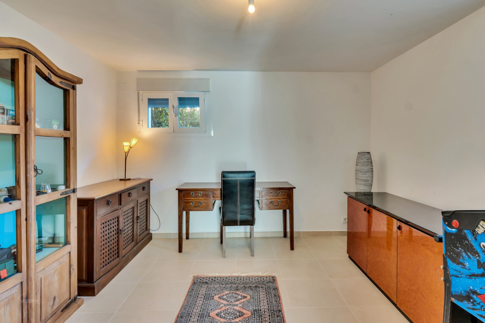 Functional home office featuring a classic wooden desk, black leather chair, and rustic cabinetry. The space includes a glass-fronted display unit and neutral tiled flooring.