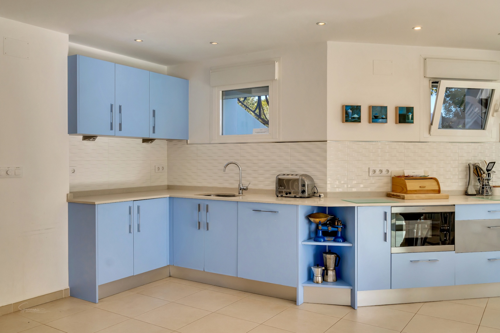 Functional kitchen with pastel blue cabinetry, light stone countertops, and a white tiled backsplash. Features integrated appliances and windows providing natural light.