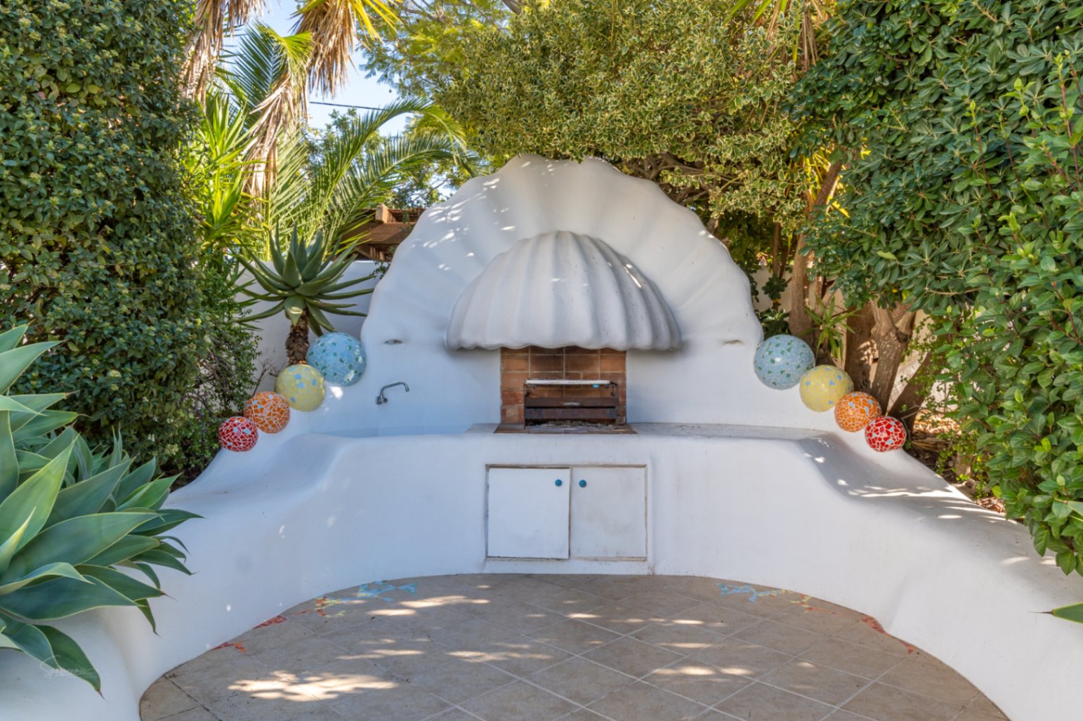 Ibiza-style outdoor BBQ area featuring a white shell-shaped hood, curved masonry counters with mosaic accents, and an integrated grill set amidst lush tropical foliage.