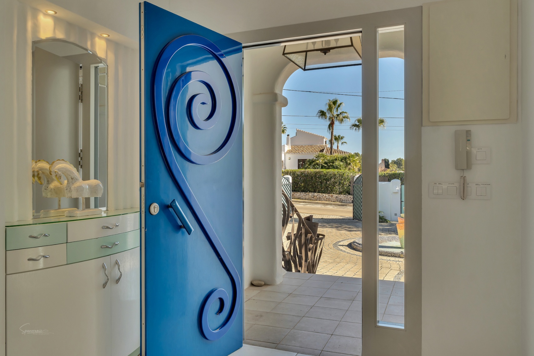 Main entrance featuring a vibrant blue door with spiral relief, leading to a bright foyer with built-in cabinetry and views of the paved garden and fountain.