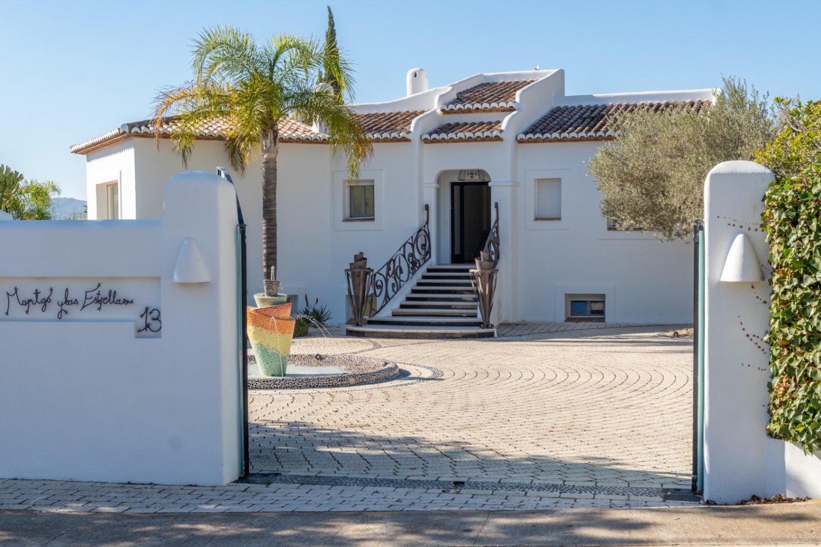Main entrance featuring a white Ibiza-style facade, ornate wrought iron railings on the stairs, and a colorful mosaic fountain in the paved courtyard.
