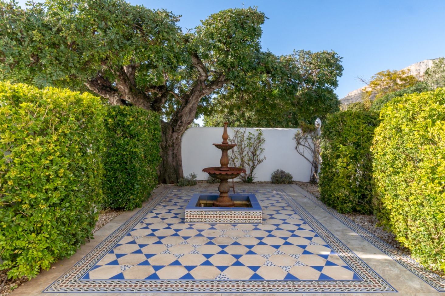 Mediterranean courtyard featuring a tiered fountain and intricate blue and white geometric tiling, framed by mature hedges and the Montgó mountain backdrop.