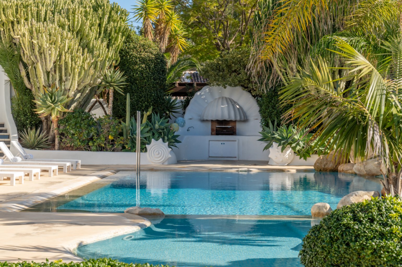 Mediterranean pool area featuring a unique shell-shaped outdoor barbecue, surrounded by lush tropical gardens, cacti, and white sun loungers.