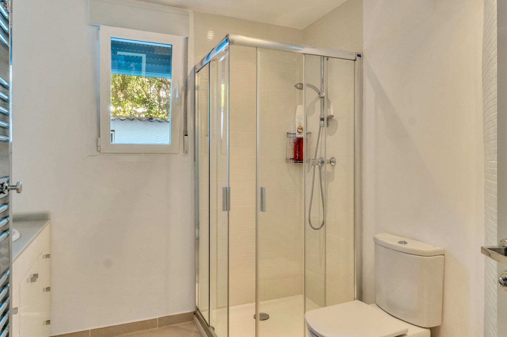 Modern bathroom featuring a glass corner shower enclosure, white vanity with storage drawers, and a window providing natural light and garden views.