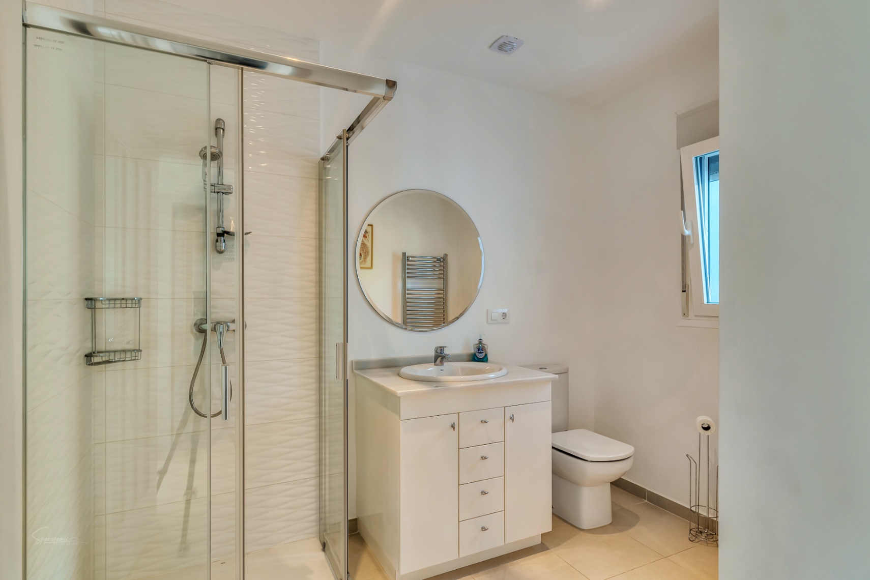 Modern bathroom featuring a glass-enclosed walk-in shower with textured tiles, a white vanity with a circular mirror, and natural light from a side window.