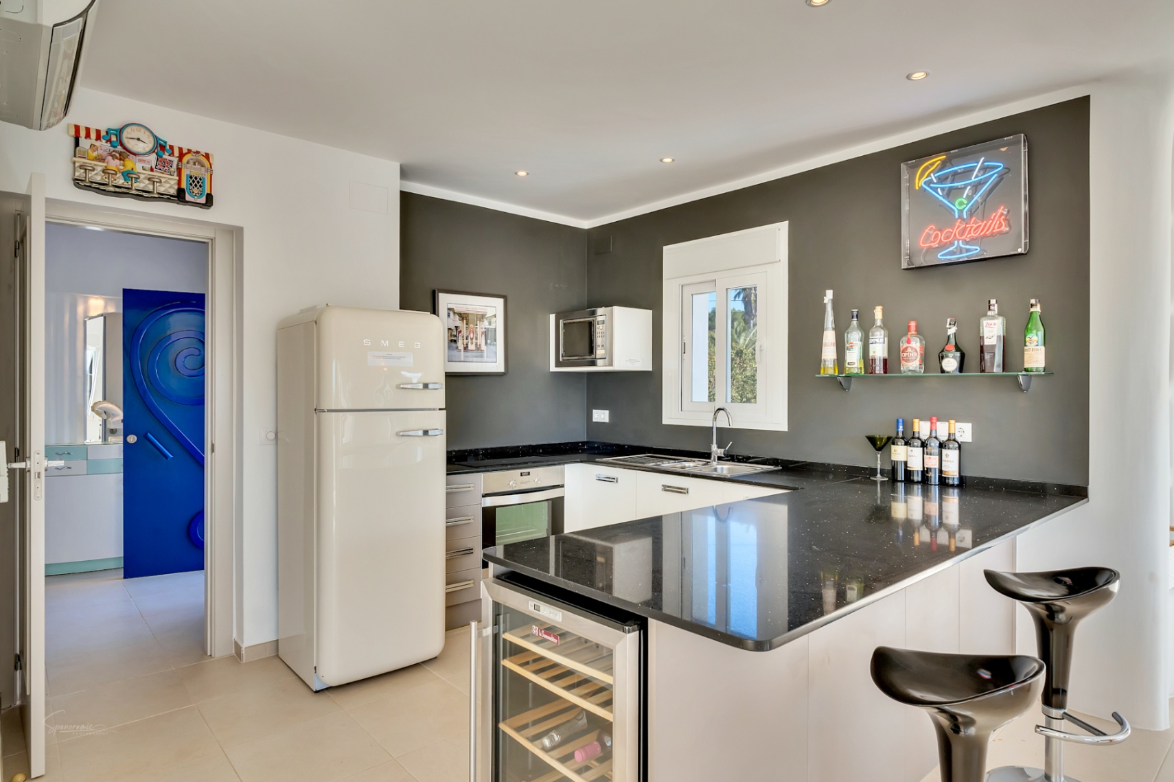 Modern kitchen featuring a black granite countertop, retro Smeg refrigerator, and a breakfast bar with an integrated wine cooler and neon cocktail signage.