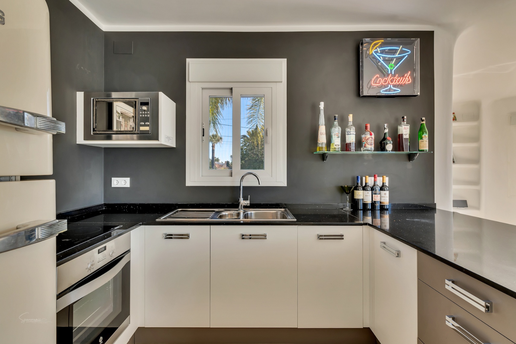 Modern kitchen featuring black countertops, white cabinetry, and a dedicated bar area with a neon sign. Includes integrated appliances and a window overlooking palm trees.