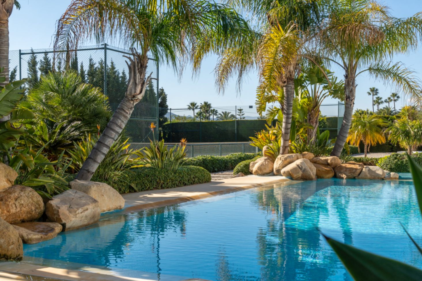 Natural-style swimming pool with rock accents and tropical landscaping, featuring a private fenced tennis court in the background.