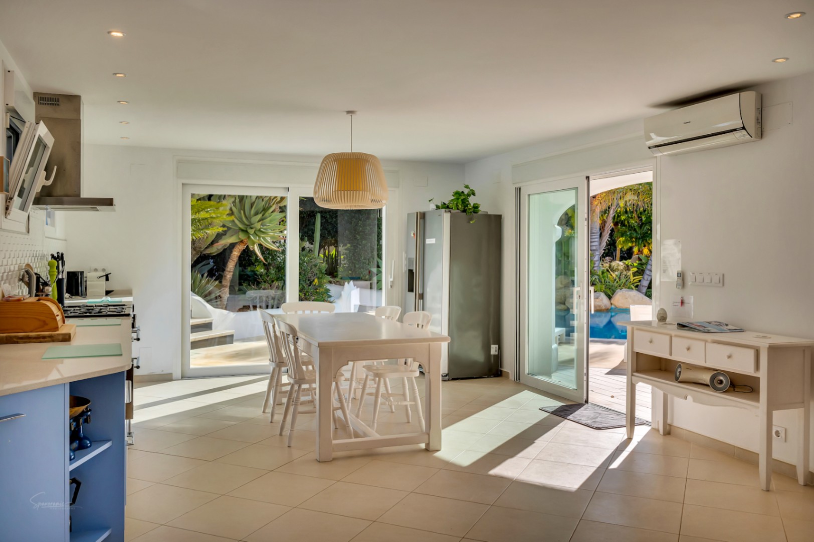Open-plan kitchen and dining area with a white table, blue cabinetry, and sliding glass doors opening to the pool and garden, featuring recessed lighting and tiled floors.