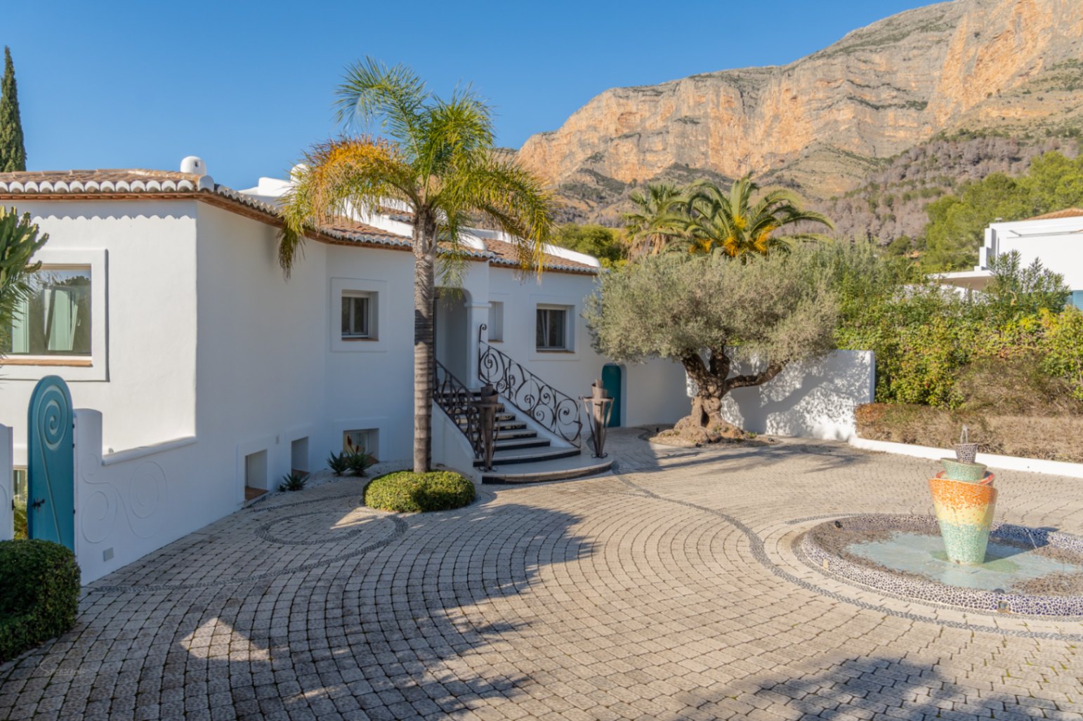Paved entrance courtyard featuring a mosaic fountain and views of the Montgó mountain. White Ibiza-style facade with wrought iron railings, palm trees, and an olive tree.