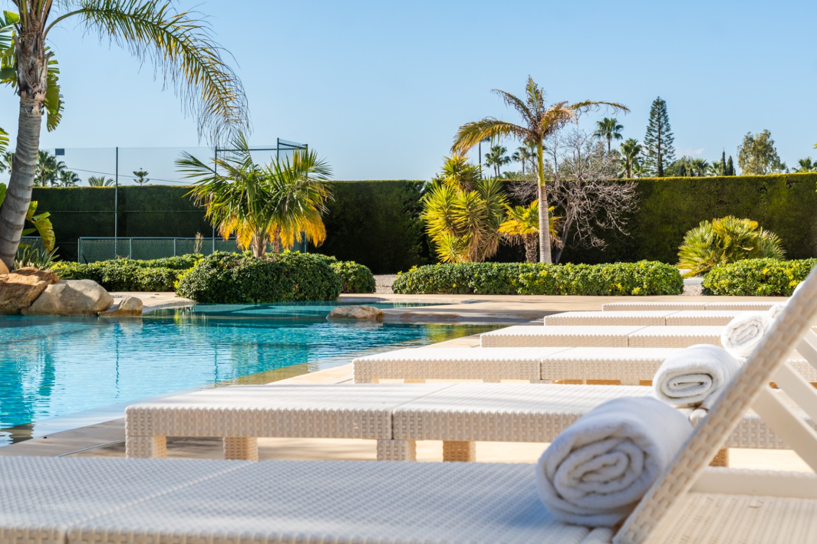 Poolside area featuring white wicker sun loungers, turquoise waters with rock accents, and a private tennis court bordered by mature landscaping.