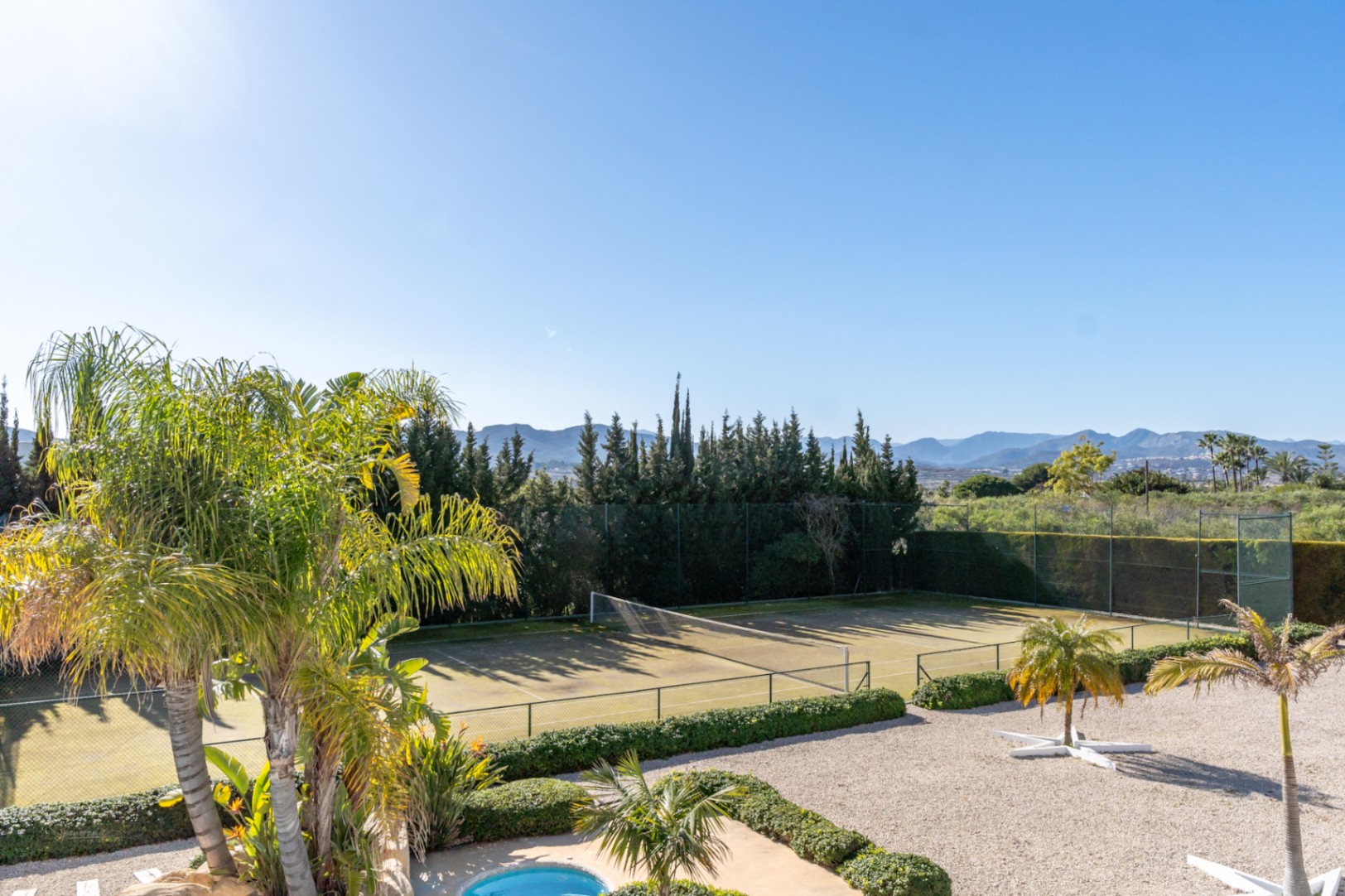Private tennis court bordered by manicured hedges and palm trees, offering expansive views of the surrounding valley and mountain range.