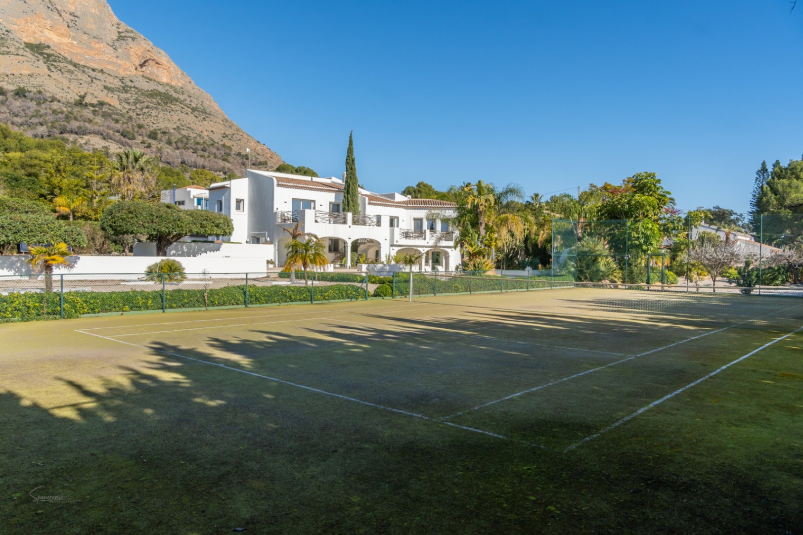 Private tennis court with a green surface and perimeter fencing, set against the white Ibiza-style villa and the majestic Montgó mountain backdrop.