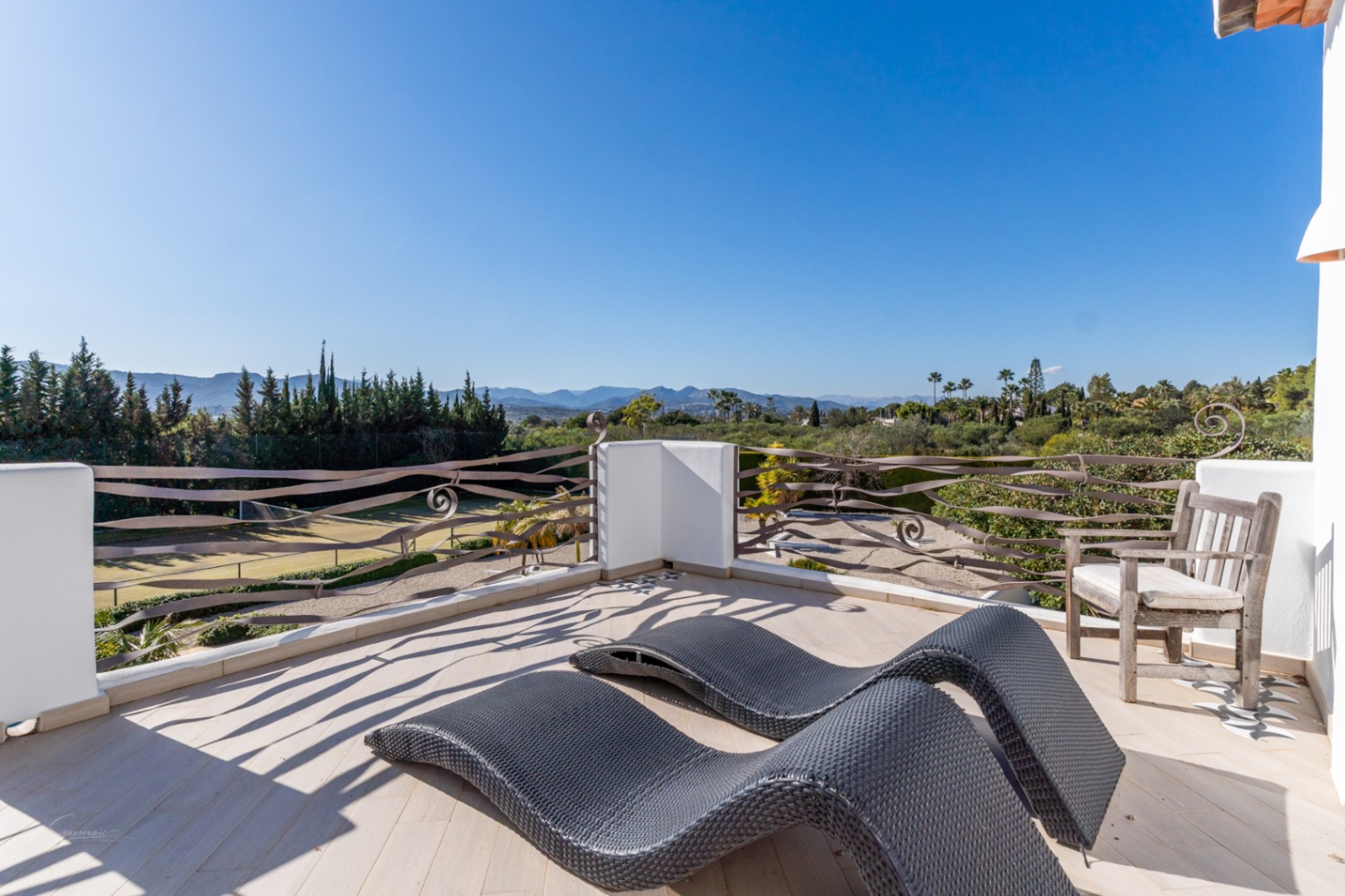 Upper terrace featuring wavy iron railings, ergonomic sun loungers, and expansive views over the private tennis court and the mountain valley.