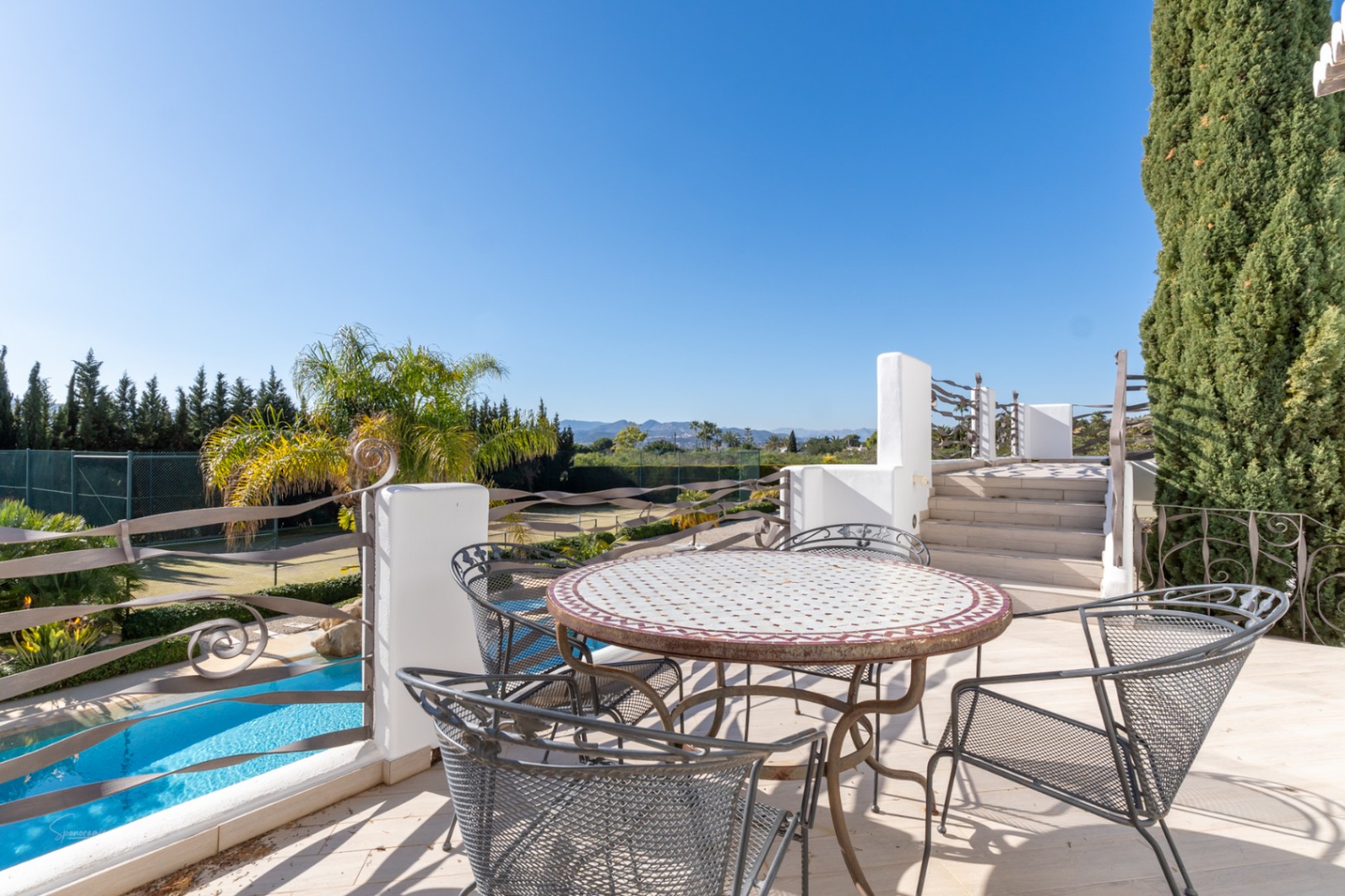Upper terrace with mosaic dining table and custom iron railings, overlooking the swimming pool, private tennis court, and mountain landscape.