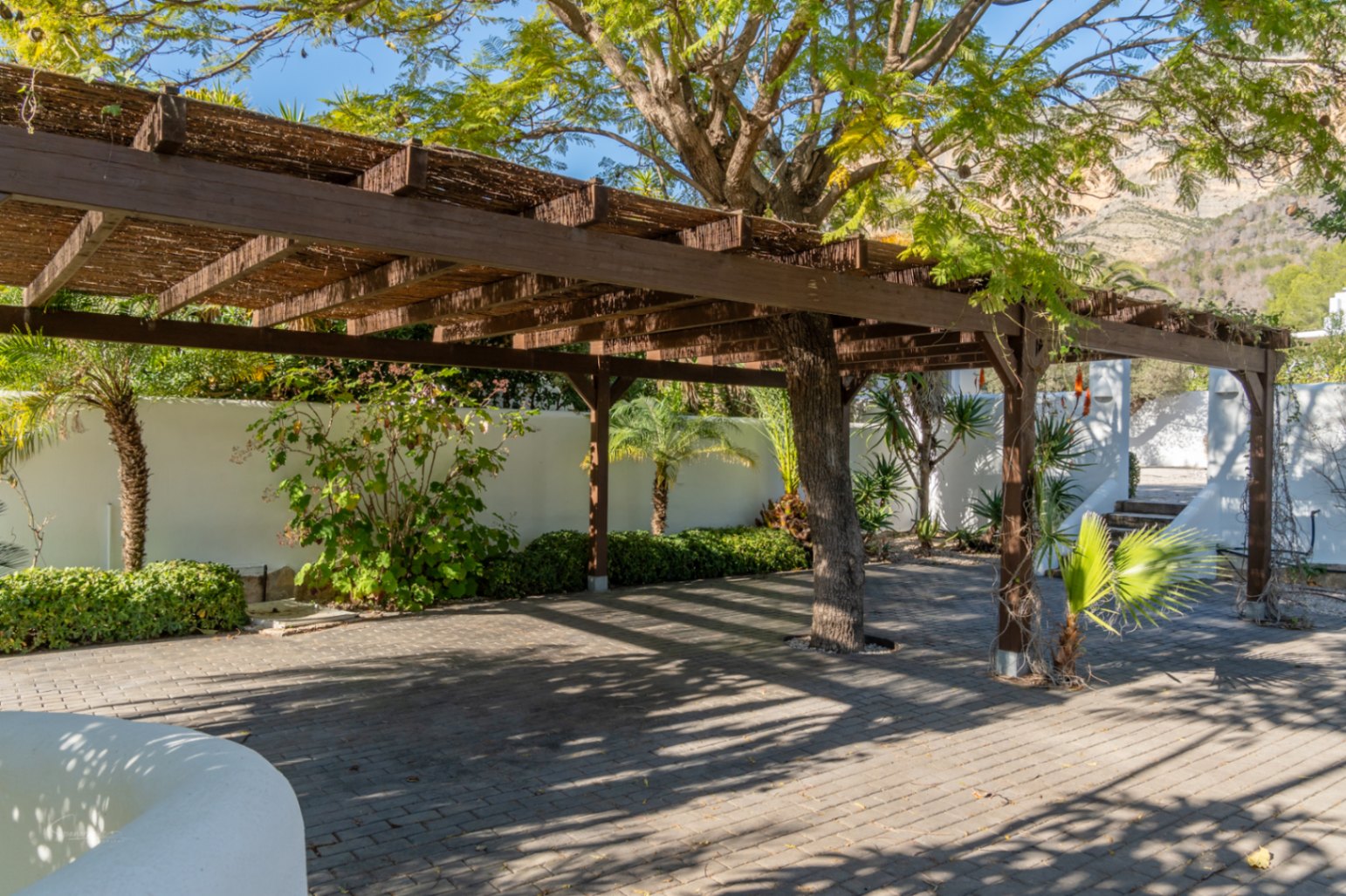 Wooden pergola with a natural reed roof integrated around a mature tree, featuring paved flooring and views of the Montgó mountain and Mediterranean gardens.
