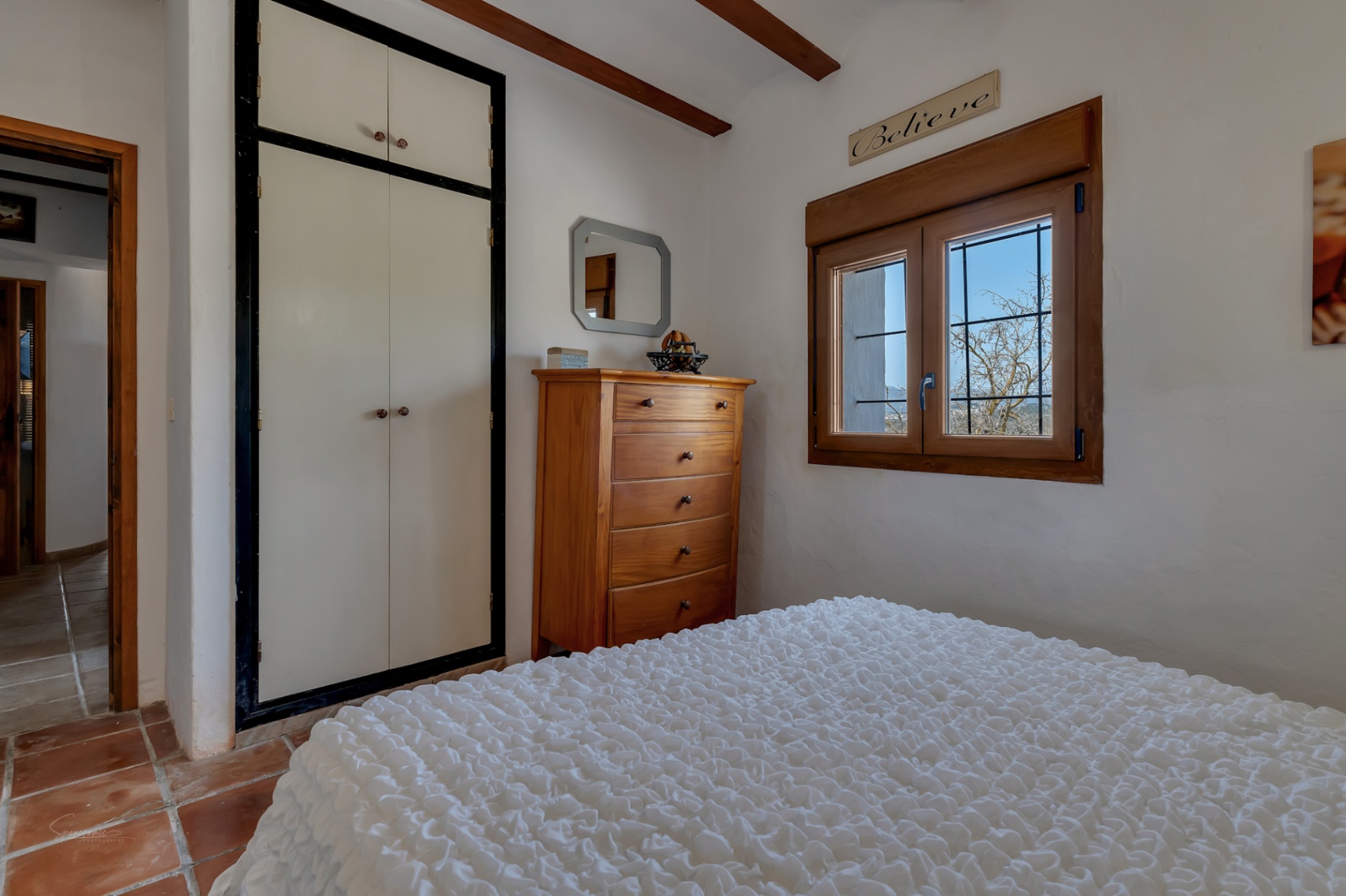 Bedroom with a white built-in wardrobe, wooden chest of drawers, and a window with wood trim.