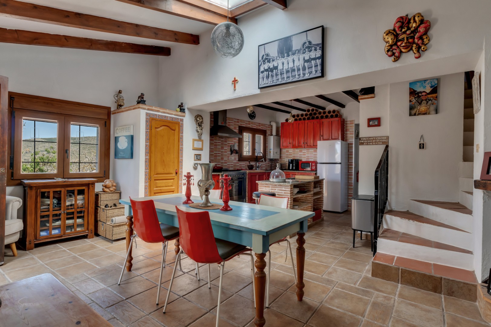 Bright dining area with a blue table, red chairs, and a red kitchen. Wooden beams and eclectic decor.