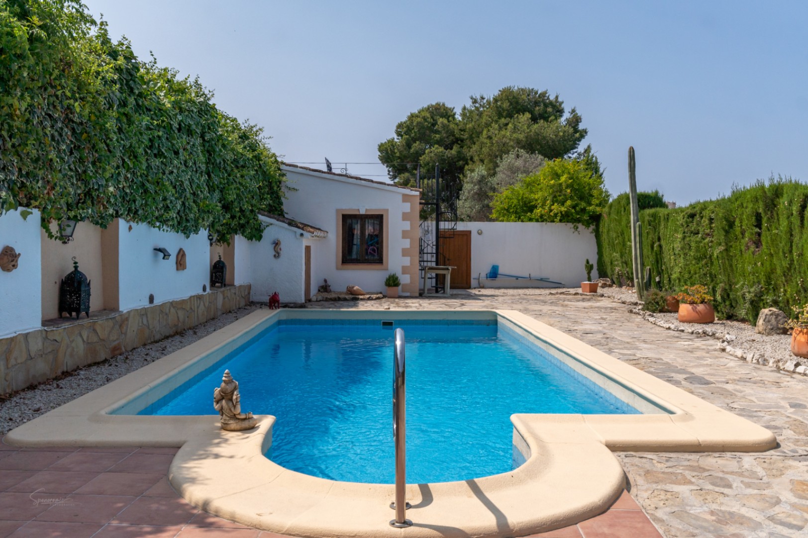 Pool view with stone trim and metal ladder, surrounded by gardens and white walls.