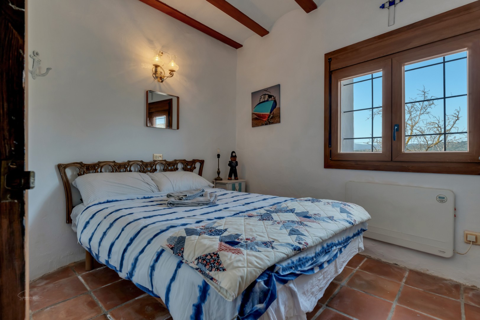 Rustic bedroom featuring terracotta flooring, a bed with a blue quilt, and a wood-framed window with outdoor views.