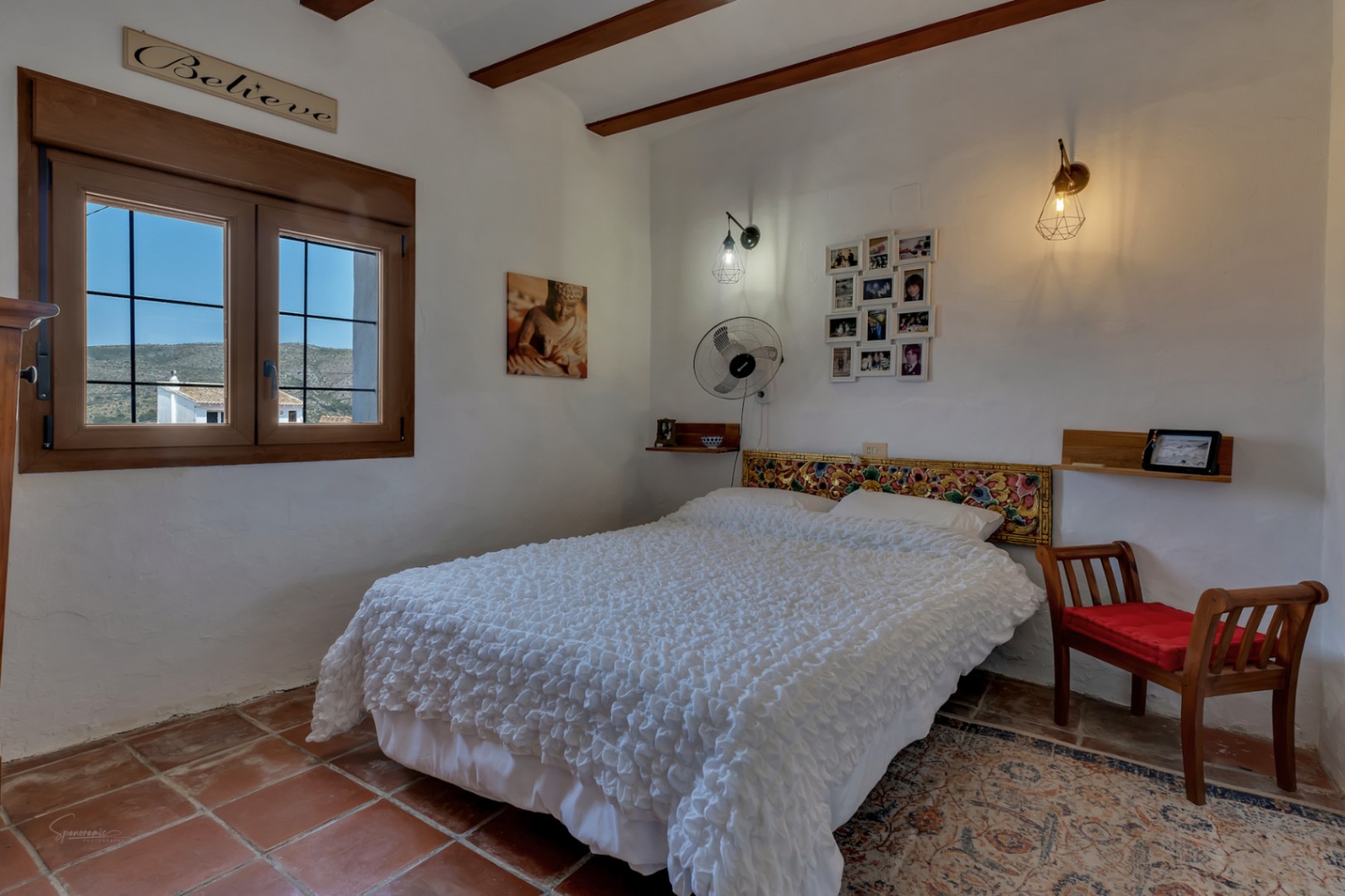 Rustic bedroom with terracotta flooring, a double bed with a carved headboard, and a window with exterior views.