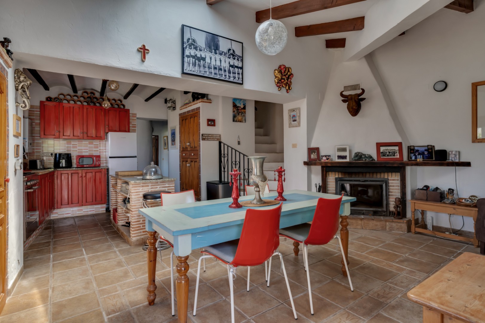 Rustic dining area with a blue table, red chairs, and a red wood kitchen. Fireplace with decor and staircase.