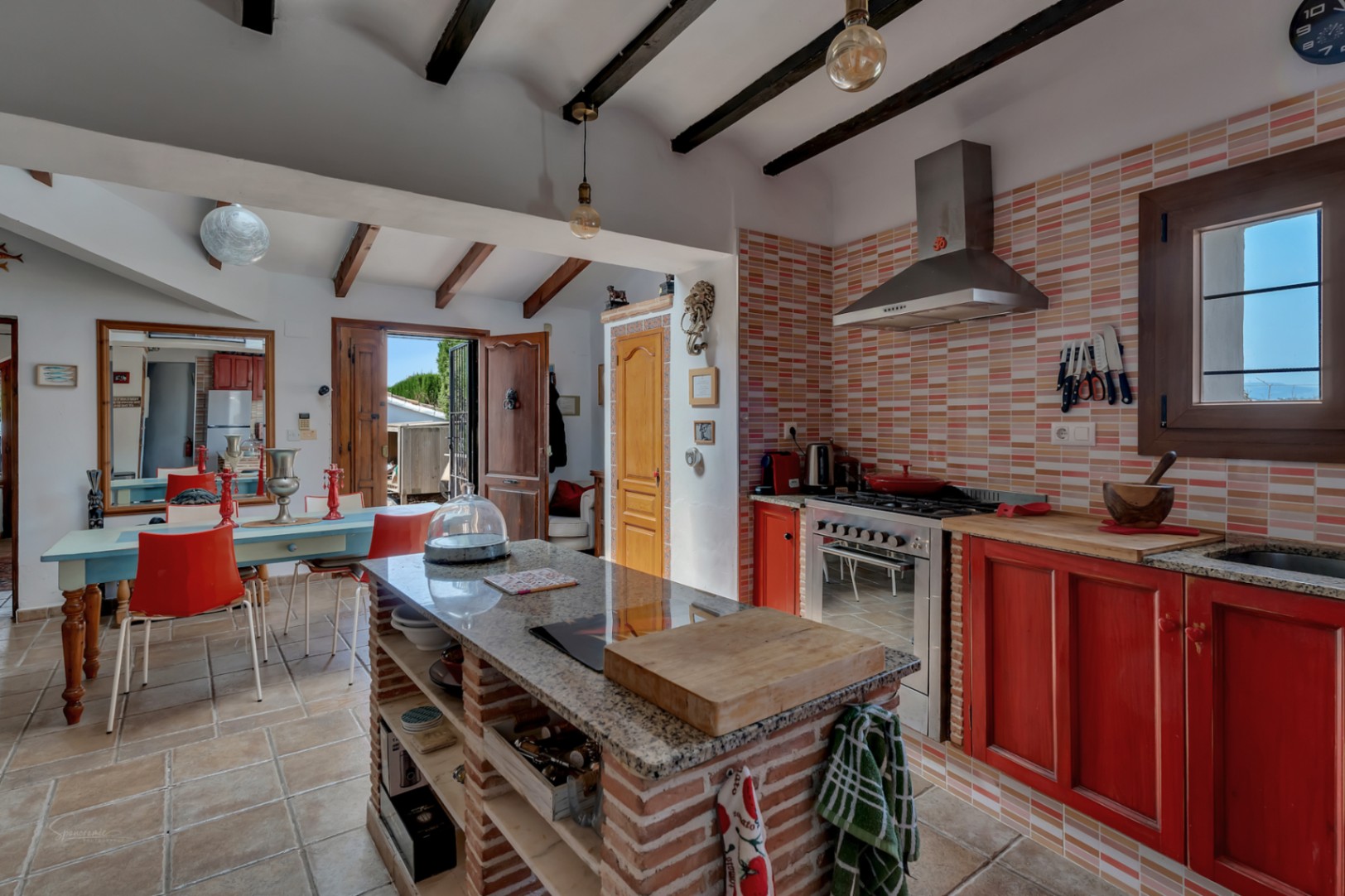 Rustic kitchen with brick island, peach-toned tile backsplash, and red cabinetry. Exposed wooden beams.