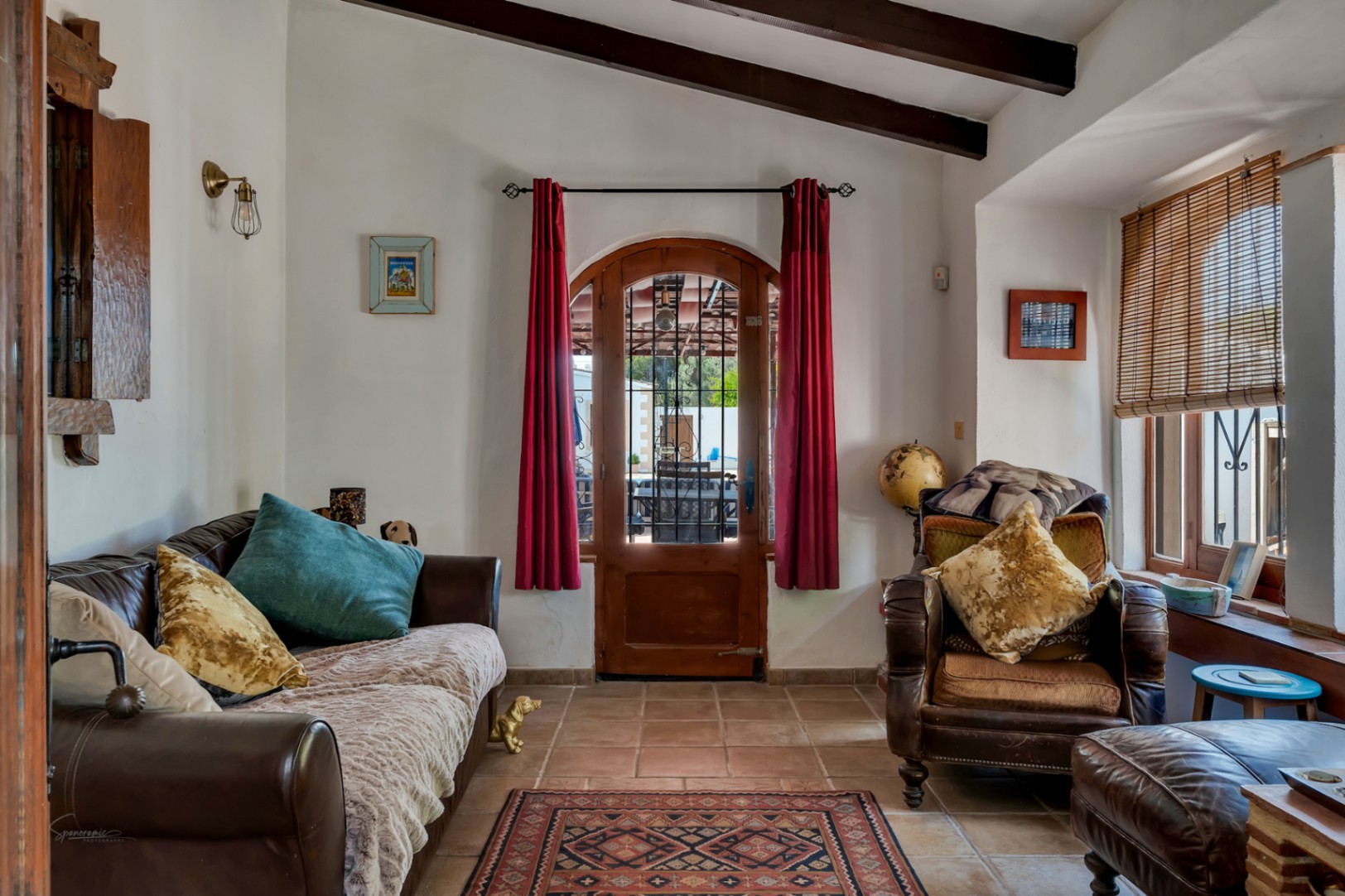 Rustic living room featuring a leather sofa, vintage armchair, and arched door with red curtains in Teulada.