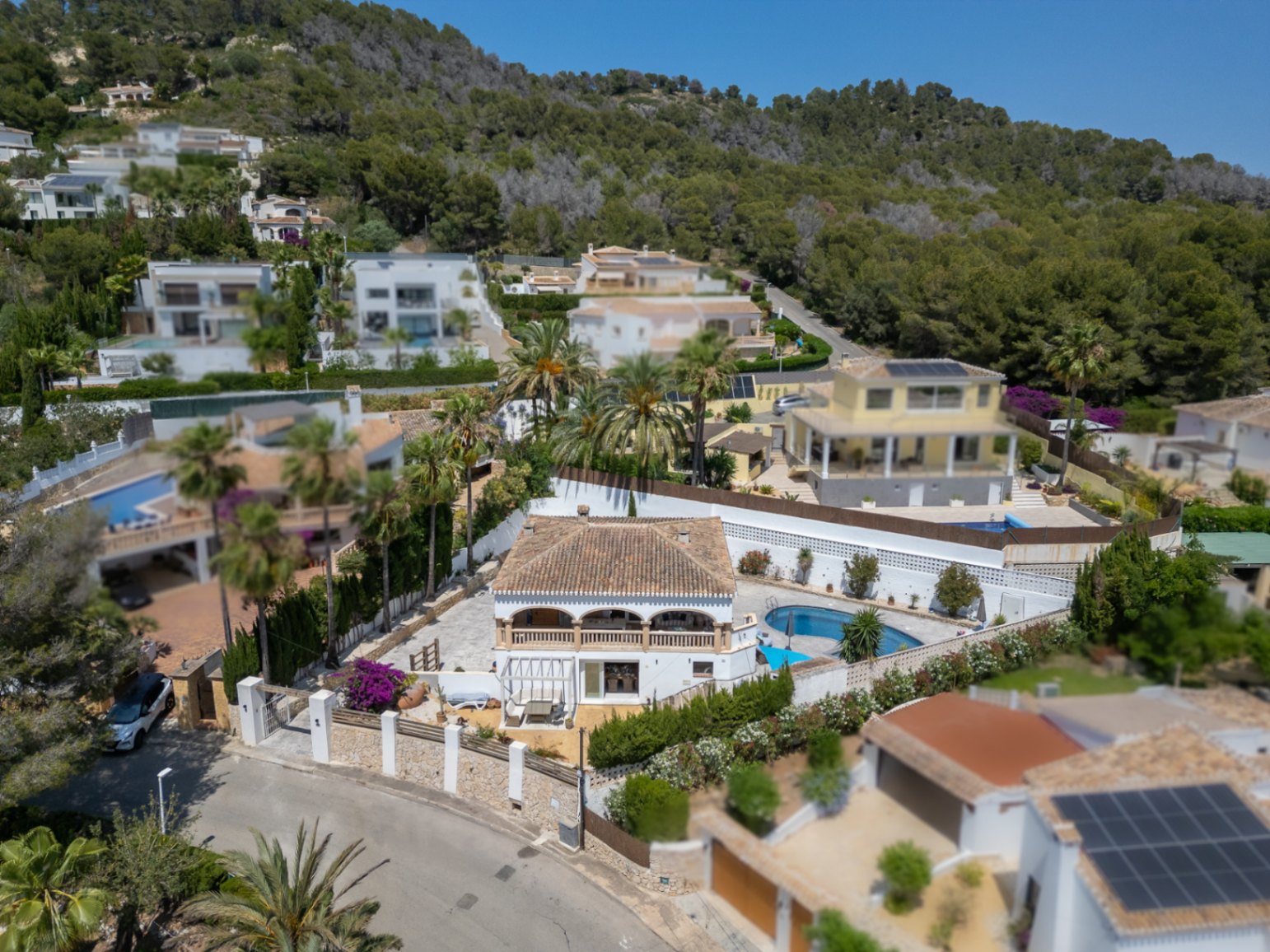 Aerial view of a Mediterranean villa with pool, surrounded by lush greenery and other residences.