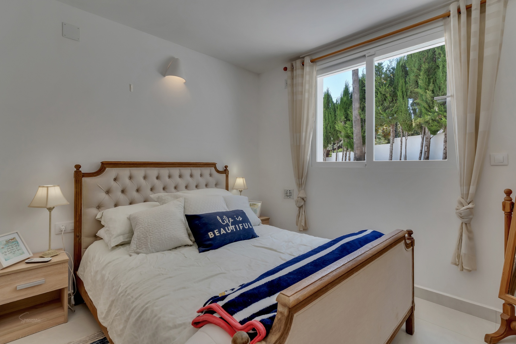 Bright bedroom featuring a wooden bed with upholstered headboard. Cypress trees visible through the window.
