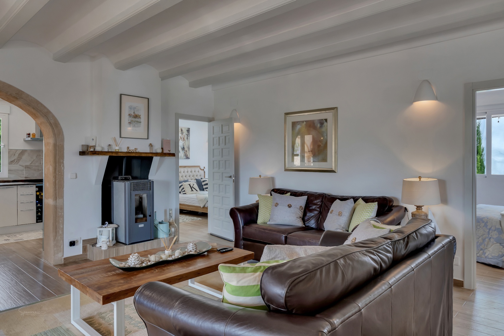 Bright living room with white beams, brown leather sofa, and modern fireplace. Views to kitchen and bedroom.