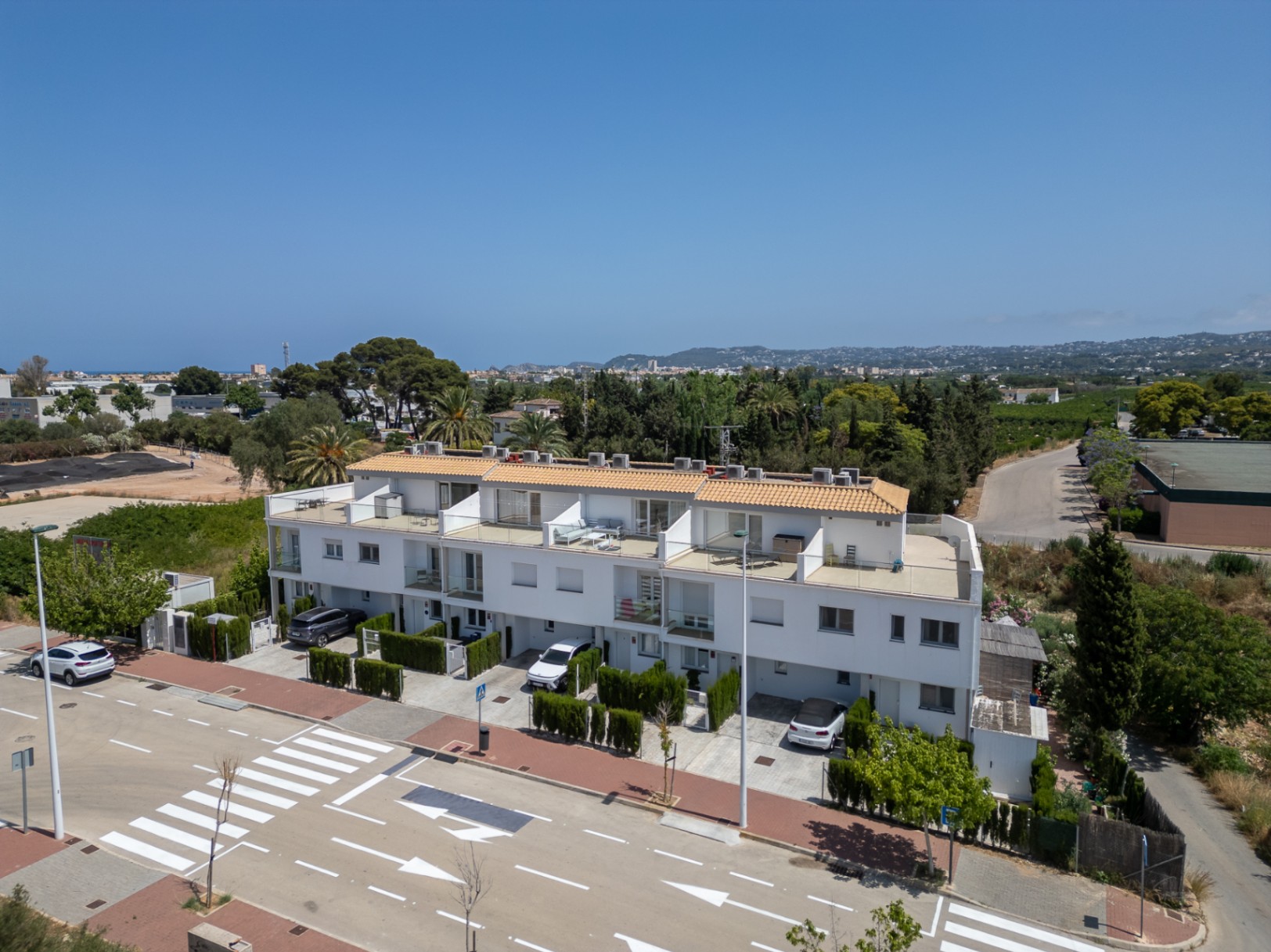 Aerial view of the modern townhouse complex featuring private parking at the entrance, white facades, and multiple levels with glass-railed terraces and tiled roofs.