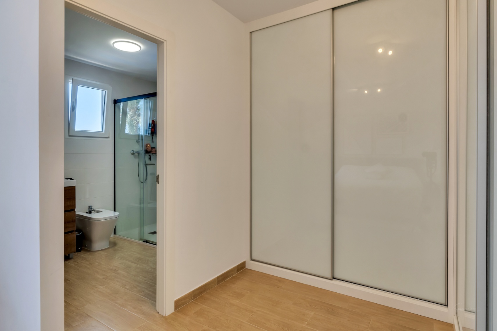 Bedroom featuring large built-in wardrobes with white sliding doors and an adjacent en-suite bathroom with a walk-in shower and wood-effect flooring.