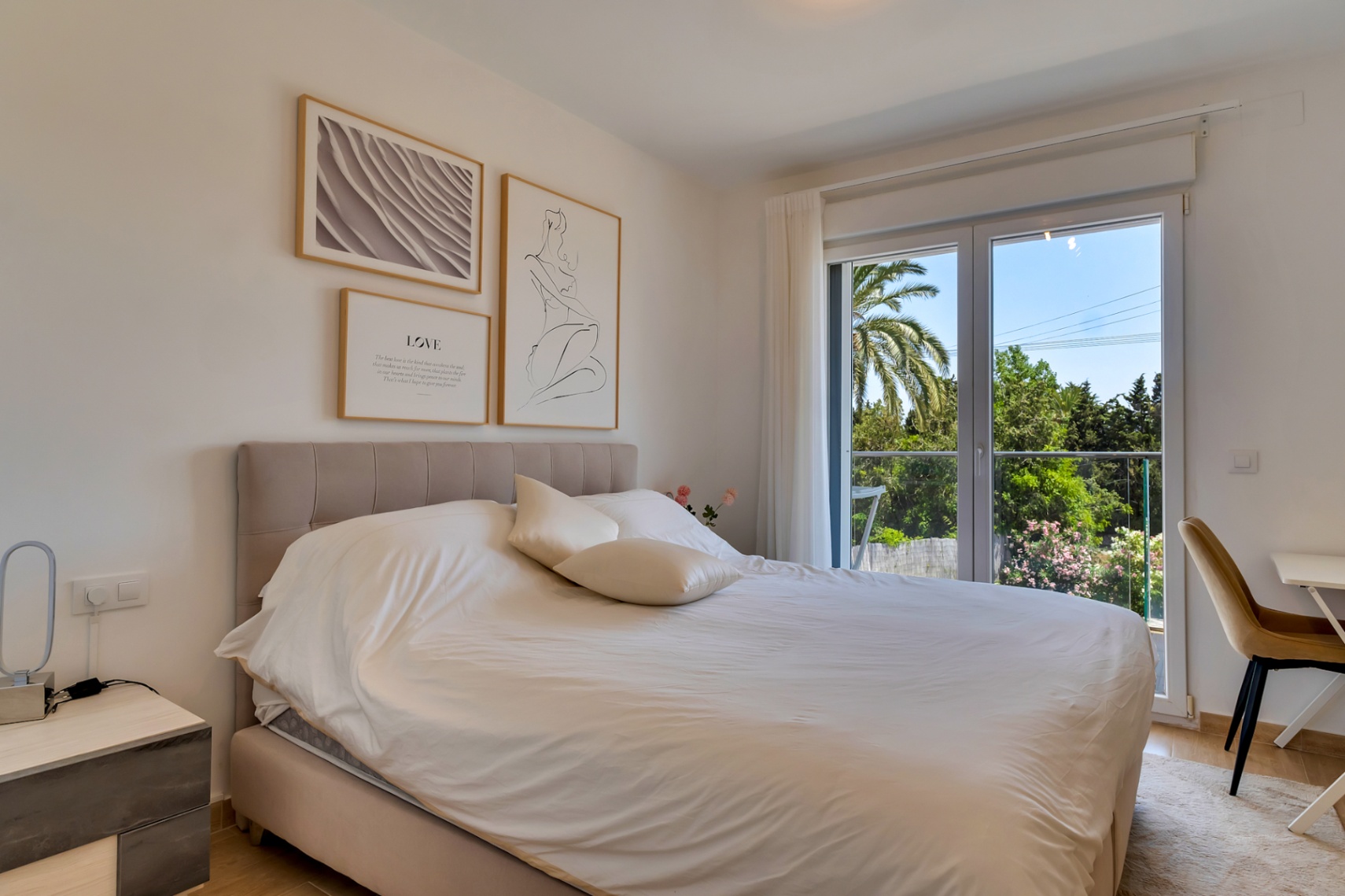 Bright bedroom featuring a double bed with a padded headboard, a dedicated workspace, and large glass doors opening to views of Mediterranean greenery and palm trees.