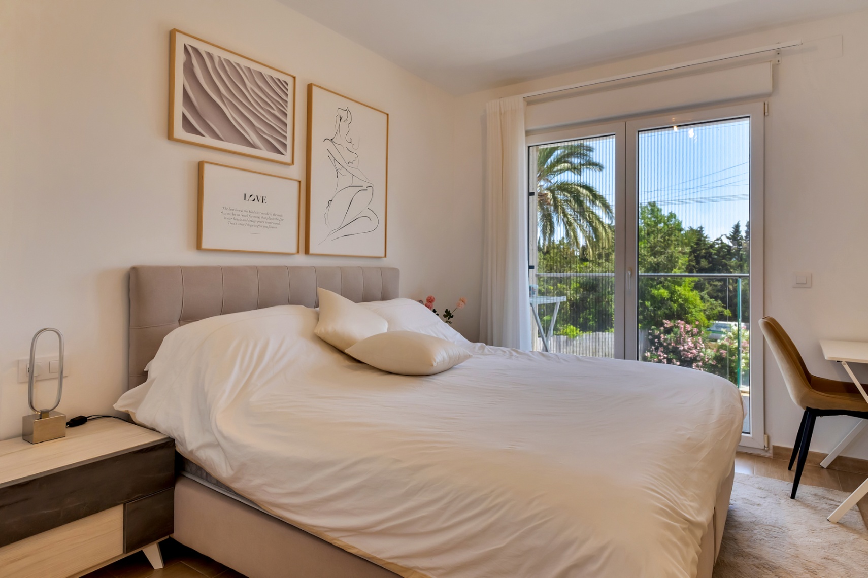 Bright bedroom featuring a double bed with upholstered headboard and a workspace. Large glass doors with mosquito screens open onto a private balcony with garden views.