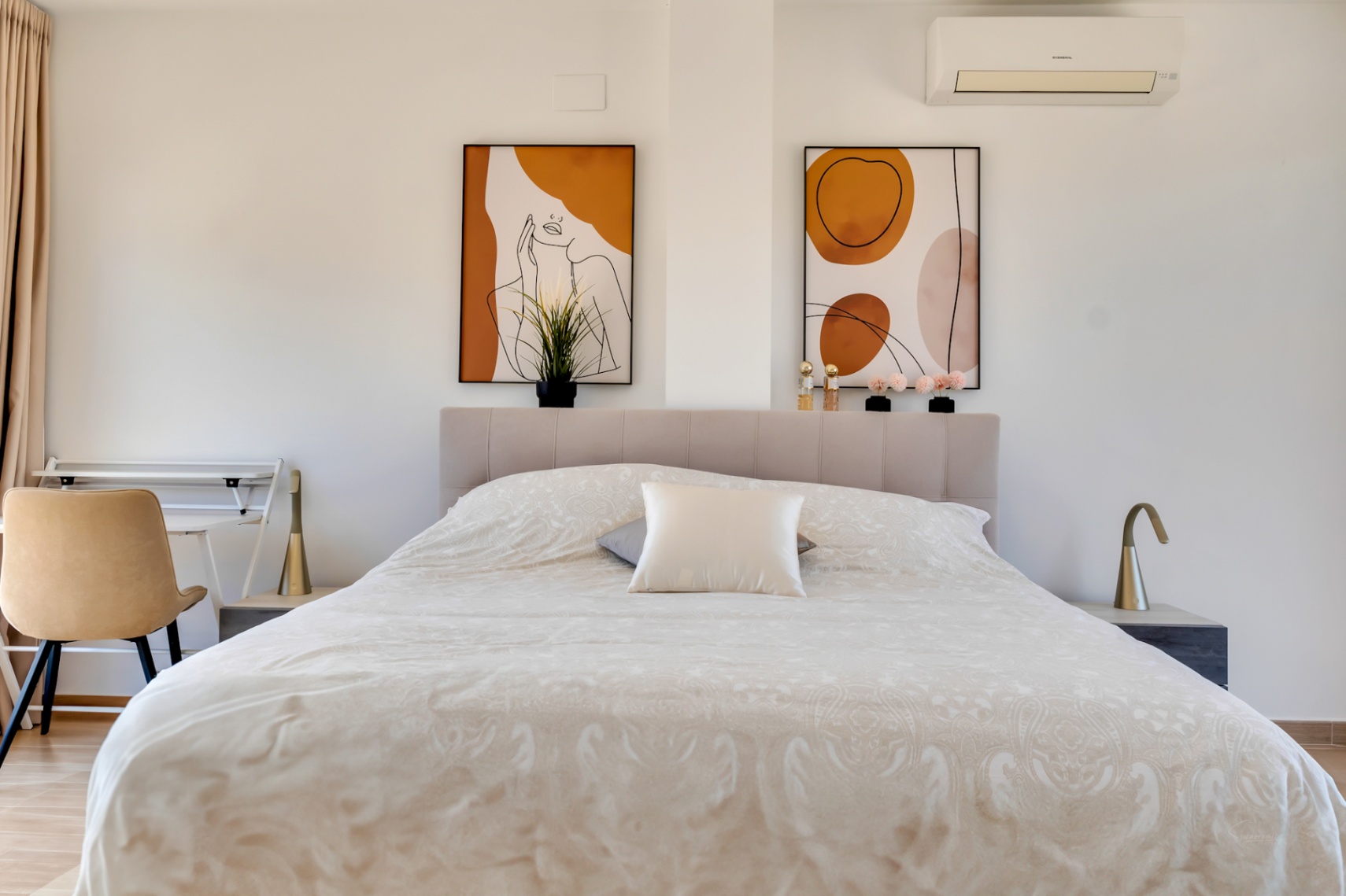 Bright bedroom featuring a tufted headboard, abstract wall art in earth tones, and a dedicated workspace. Equipped with a wall-mounted air conditioning unit.