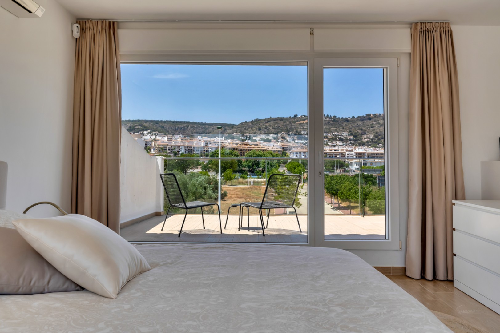 Bright bedroom featuring direct access to a private terrace with outdoor seating. Large glass doors frame panoramic views of the town and surrounding hills.
