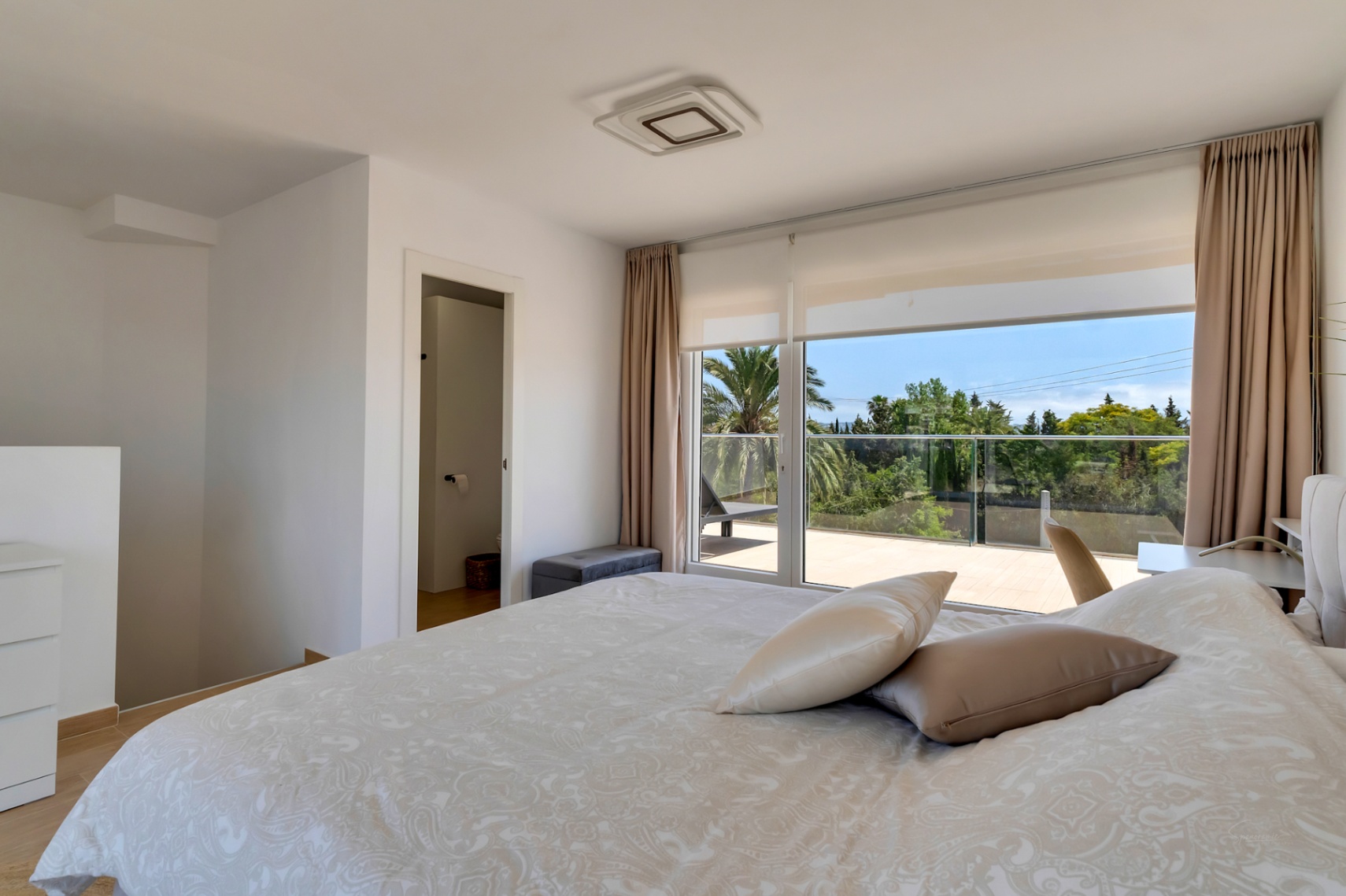 Bright bedroom featuring large glass doors opening onto a private terrace with green views. Includes integrated air conditioning and ensuite bathroom access.