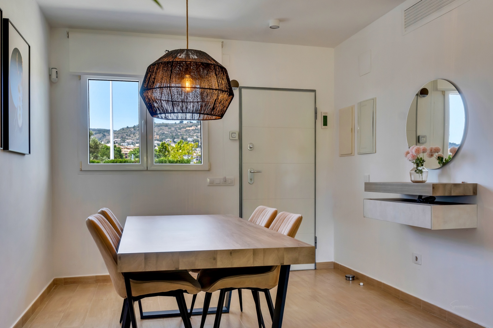 Bright dining area featuring a solid wood table, natural fiber pendant light, and hillside views. Includes a modern floating console and circular mirror.