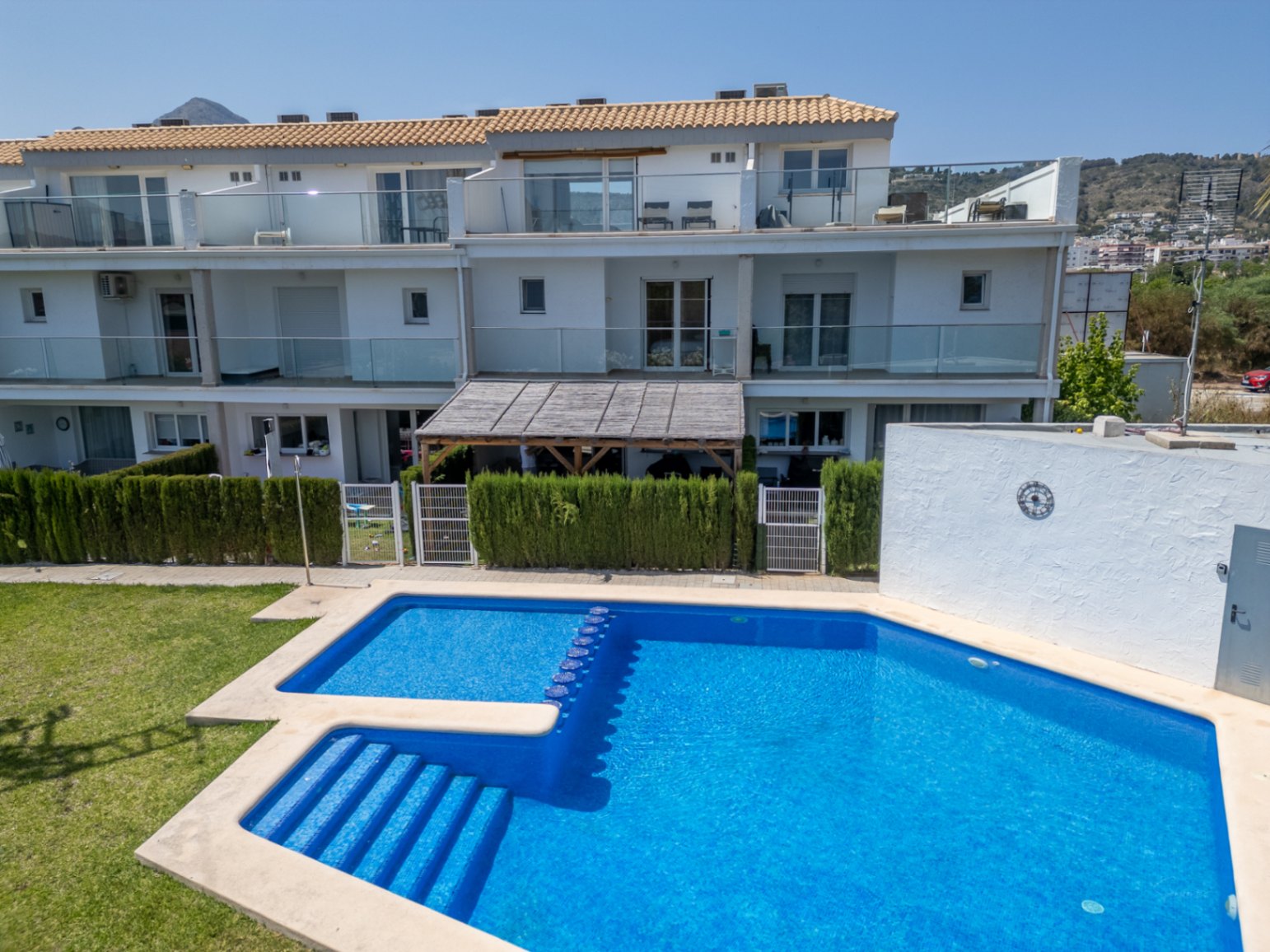 Communal swimming pool with blue tiling and integrated steps, set before the modern white facade featuring glass balconies and private patios with wooden pergolas.