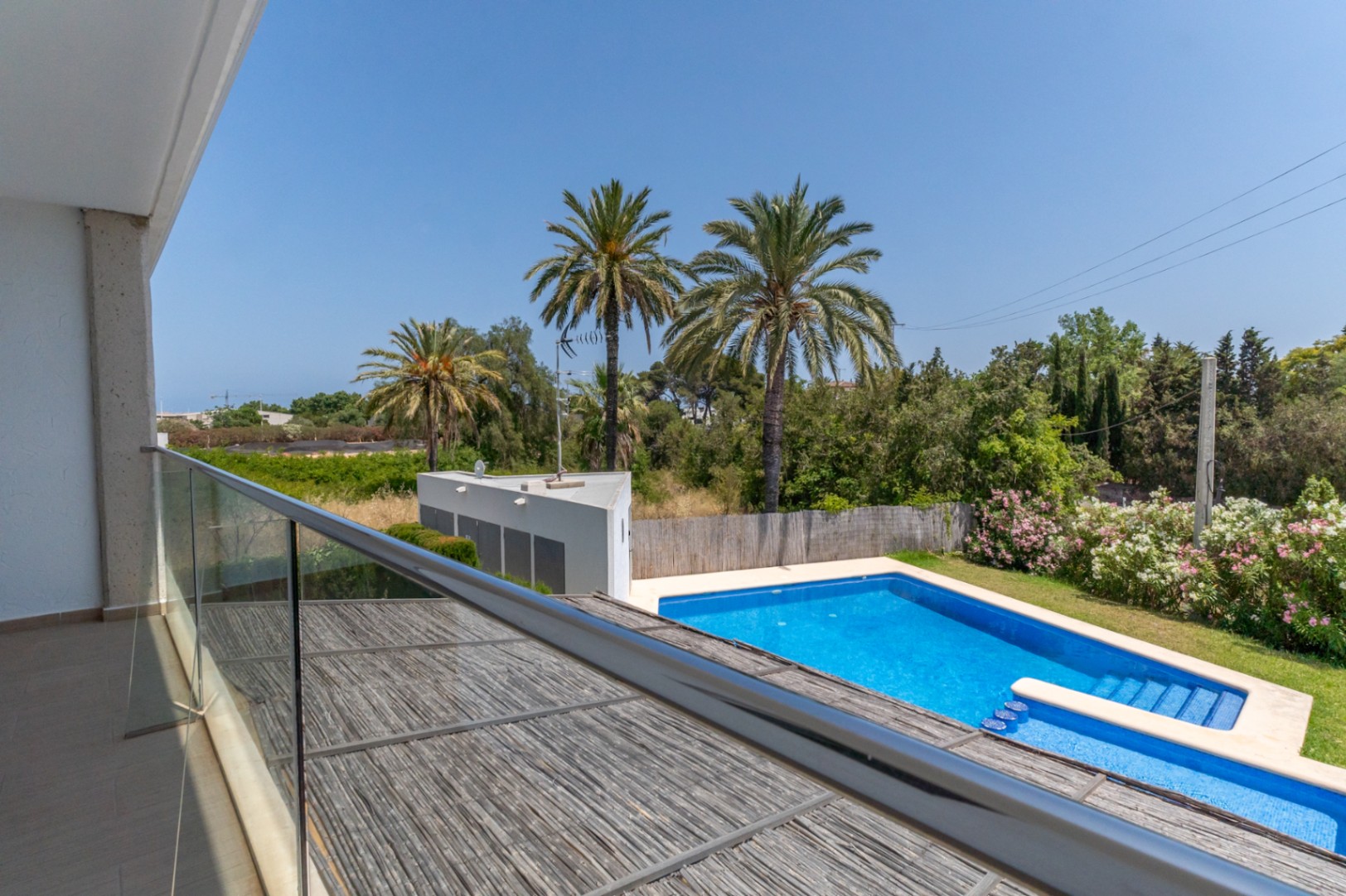 Elevated view from the upper terrace featuring a glass balustrade, overlooking the communal pool, Mediterranean gardens with palms, and the private patio's pergola.