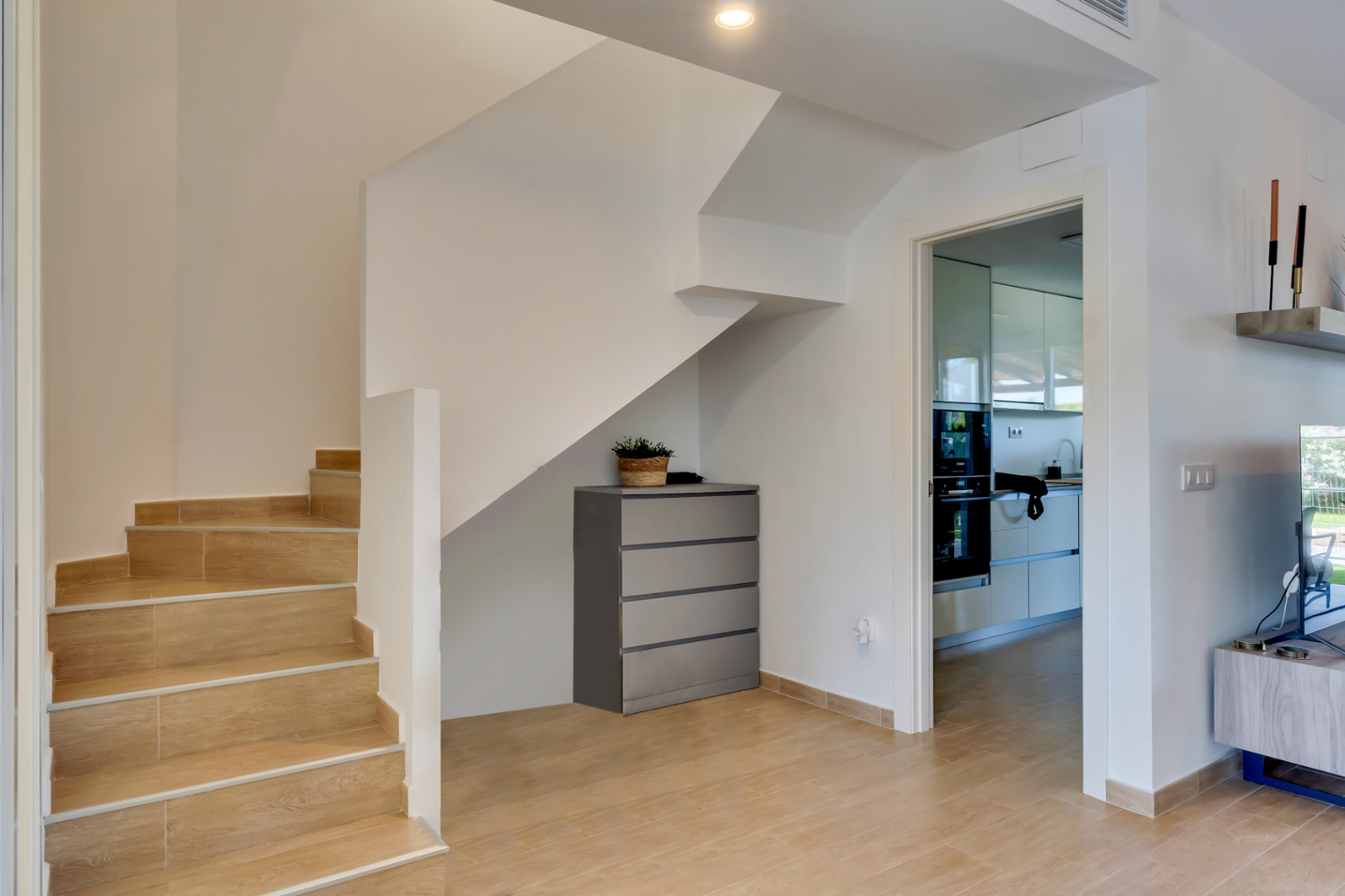 Interior staircase featuring wood-effect ceramic tiles and white walls. The view shows the transition to the modern kitchen and a functional storage area under the stairs.