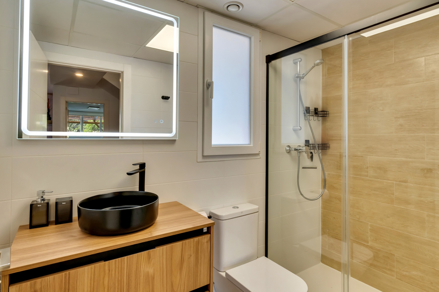 Modern bathroom featuring a black vessel sink on a wood vanity, backlit LED mirror, and a walk-in shower with black framing and wood-effect tiling.