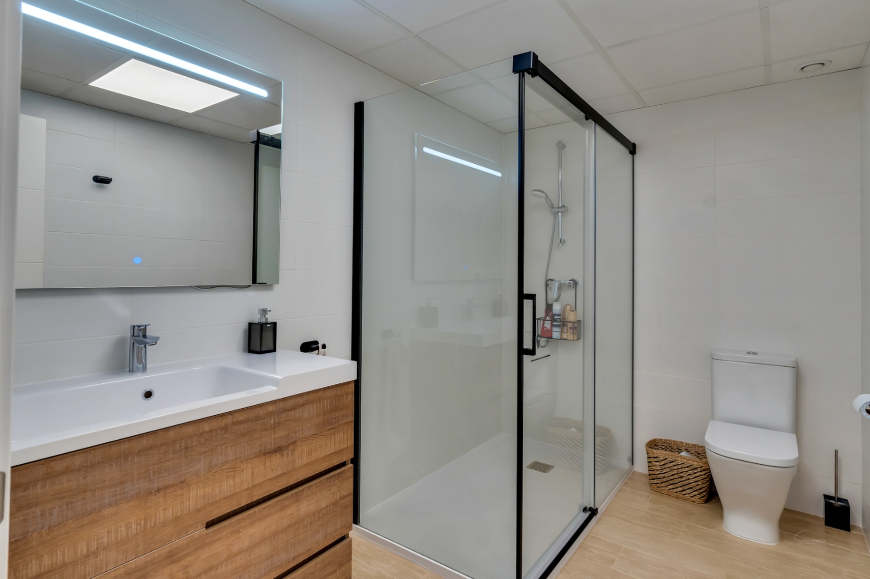 Modern bathroom featuring a wood-effect vanity, integrated sink, and LED-lit mirror. It includes a spacious walk-in shower with black framing and wood-look floor tiles.